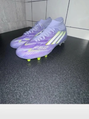 Lila Adidas F50 fotbollsskor - Säljer ett par snygga Adidas F50 fotbollsskor i lila med vita och neongula detaljer. Skorna har hög krage för extra stöd och snörning framtill. Yttermaterialet är syntetiskt och dobbarna är anpassade för spel på gräs.  Storlek 36 1/3