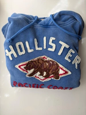 Blå Hollister hoodie med björn - Säljer en blå hoodie från Hollister med broderad björn och texten 'Pacific Coast' framtill. Hoodien har känguruficka, dragsko i huvan och ribbade muddar. Perfekt för dig som gillar en avslappnad och sportig stil. Knappt använd iom för liten för mig.