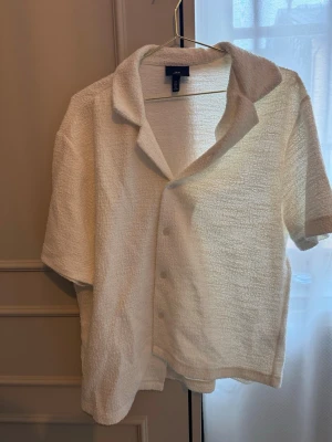 Vit frottéskjorta från H&M - Säljer en vit skjorta från H&M i frotté med kort ärm och klassisk krage. Skjortan har knappar framtill och en avslappnad passform, perfekt för en chill och trendig look.