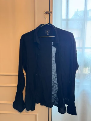 Svart skjorta från H&M, regular fit - Svart långärmad skjorta från H&M i regular fit. Skjortan har klassisk krage, knäppning framtill och är tillverkad i ett lätt, något transparent tyg som ger en avslappnad vibe. Perfekt att styla lager på lager eller bära som den är.