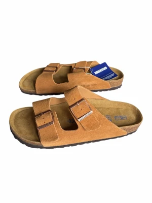 Birkenstocks brun - Snygga sandaler från Birkenstock i brun mocka med två justerbara remmar och metallspännen. Fotbädden är ergonomiskt utformad och sulan är i kork och gummi för extra komfort. Perfekta för en avslappnad och trendig stil. Aldrig använts 
