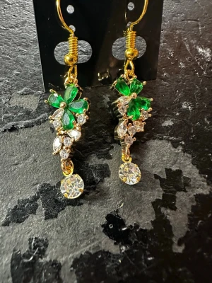 Guldiga örhängen med gröna stenar - Säljer ett par eleganta örhängen i guldfärg med gnistrande gröna och klara stenar. Örhängena har en blommig design med bladformade detaljer och en rund klar sten som hänger längst ner. Perfekta för dig som vill sticka ut med färg och bling.