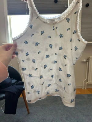 Vit blommig linnetopp från Lindex - Söt vit linnetopp från Lindex med små blå blommor och spetsdetaljer längs kanterna. Toppen har en liten rosett framtill och är ärmlös, perfekt för varma dagar. Mjuk och bekväm bomullskvalitet.