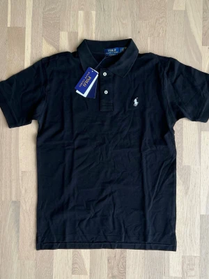 Ralph Lauren Black Polo shirt size M - Brand new, never worn Polo Ralph Lauren Custom Slim Fit polo shirt in black. Comes with original tags attached and original packaging.  * Condition: Brand New with Tags (NWT) * Color: Black * Fit: Custom Slim Fit * Size: M * Material: 100% Cotton * No flaws, stains, holes, or signs of wear. * Authentic Polo Ralph Lauren. * Retail ≈ 180$ A timeless wardrobe essential, perfect for both casual and smart-casual outfits. 