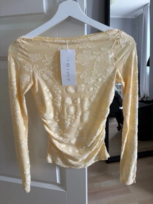 Gul blommig spetsblus  - Supersöt gul långärmad blus från New yorker/Amisu med blommigt spetsmönster. Toppen har en figurnära passform och är tillverkad i ett tunt, genomskinligt material med fina broderade detaljer. Perfekt för dig som vill ha en romantisk och trendig look till sommarn och våren. 