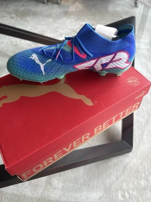 Blåa Puma fotbollsskor med neon detaljer - Säljer ett par snygga blåa Puma fotbollsskor med mintgröna dobbar och coola neonrosa och vita detaljer. Skorna har stickad ovandel för bra passform och stor FR-logga på sidan. Perfekta för dig som vill sticka ut på planen! Skorna är helt nya köpte två par men kunde ej skicka tillbaks ena så dessa har ej använts någon gång. Pris kan diskuteras!