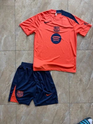 Barça träningsset orange/marinblå Nike - Säljer ett FC Barcelona träningsset från Nike med orange t-shirt och marinblå shorts. Tröjan har klubbmärke och sponsorlogga framtill samt marinblå detalj på axeln. Shortsen är marinblå med klubbmärke och Nike-logga. Perfekt för fotbollsträning eller sport.