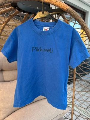 Blå Pikkuveli t-shirt Fruit of the Loom - Blå t-shirt från Fruit of the Loom med texten 'Pikkuveli' tryckt på bröstet. Klassisk rund hals och korta ärmar. Tillverkad i mjuk bomull, perfekt för en avslappnad och enkel stil.