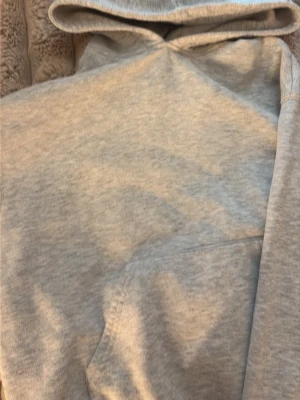 Ljusgrå hoodie med ficka - En basic ljusgrå hoodie med huva och magficka. Hoodien har ribbade muddar vid ärmslut och nederkant. Perfekt för en chill och avslappnad stil. Materialet ser ut att vara mjukt och skönt, troligen bomullsblandning.