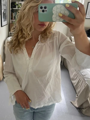 Vit blus med knappar - Säljer en vit, långärmad blus i tunn och luftig bomull. Blusen har v-ringning med knappar framtill och lätt puffade axlar. Ärmarna är vida med knytband vid ärmsluten.