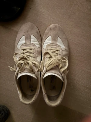 Beige och vita sneakers i mocka och läder - Säljer ett par stilrena sneakers i beige mocka och vitt läder med klassisk gummisula. Skorna har vita snören och en låg silhuett som passar till många olika outfits. Perfekt för dig som gillar minimalistisk och tidlös design.