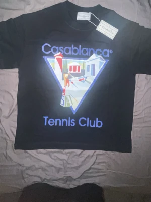 Casablanca Tennis Club t-shirt black  - Svart t-shirt från Casablanca med stort färgglatt tryck framtill där det står 'Casablanca Tennis Club' och ett motiv av tennisbana och byggnader. Klassisk passform och rund hals. Perfekt för dig som gillar streetwear med sportig vibe.