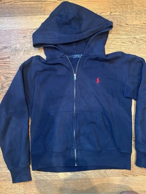 Mörkblå hoodie från Polo Ralph Lauren - Mörkblå hoodie med dragkedja från Polo Ralph Lauren. Klassisk design med röd broderad logga på bröstet, stor huva och kängurufickor framtill. Tillverkad i mjukt bomullsmaterial som känns skönt mot huden. Perfekt för en avslappnad och stilren look. Köpt på NK! Köptes ny för runt 3000kr! Men priset som jag har lagt är ej fast! Så det är frittfram att komma med bud! Tveka inte på atg höra av er!