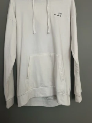 Vit hoodie med tryck EYE ALL EYE - Säljer en vit hoodie med huva och dragsnören. På bröstet finns ett litet svart tryck med texten 'EYE ALL EYE' och på huvan står det 'EYE'. Hoodien har en stor magficka och långa ärmar. Perfekt för en clean och enkel stil.
