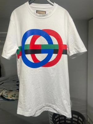 Vit Gucci t-shirt med färgglad logga - Vit t-shirt från Gucci med stor, färgglad logga framtill i blått, rött, grönt och svart. Klassisk rund halsringning och korta ärmar. Tillverkad i mjuk bomull för en skön känsla och stilren look.                               Storlek L men passar mig som s helt ok och passar förmodligen m bra också.