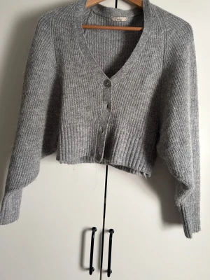 Grå ribbstickad kofta med knappar - Mysig grå kofta i ribbstickat material med v-ringning och knäppning framtill. Kortare modell med långa ärmar och breda muddar. Perfekt för lager på lager och enkel att styla till olika outfits.