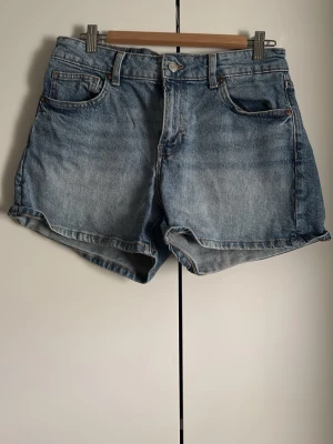 Blå denimshorts med fem fickor - Klassiska blå jeansshorts i denim med fem fickor och uppvikt kant. De har en relaxed passform och stängs med knapp och dragkedja. Perfekta för varma dagar och enkla att matcha med olika stilar.