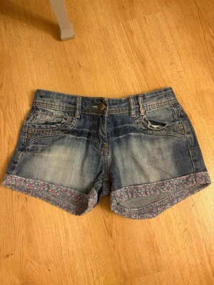 Blå jeansshorts med blommiga detaljer - Säljer ett par blå jeansshorts med uppvikta benslut som har ett blommigt mönster på insidan.  Shortsen är lågmidjade och har klassiska fickor fram och bak, bälteshällor och knappstängning. Dessutom finns det justerbara knappar för midjan på insidan. Står tyvärr ingen storlek men gissar på XS eller XXS.
