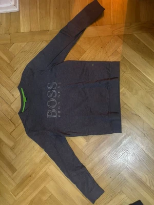 Grå sweatshirt från Hugo Boss - Mörkgrå sweatshirt från Hugo Boss med stort BOSS-tryck och broderad logga på bröstet. Tröjan har rund halsringning och långa ärmar. Insidan av kragen har en grön detalj som ger en snygg kontrast. Perfekt för en avslappnad och stilren look.