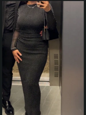 Svart lång prickig meshklänning - Snygg svart långklänning med vita små prickar och meshdetaljer på ärmarna. Figurnära passform och rund halsringning. Perfekt för dig som vill ha en elegant och trendig look. Klänningen har långa ärmar och transparent mesh över armarna. Ny pris 699kr
