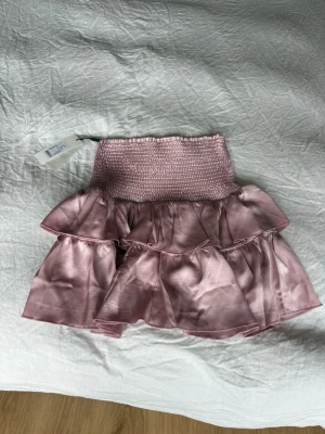 Rosa volangkjol i satin från Neo Noir - Supersöt kort kjol i satin från Neo Noir i en mjuk rosa nyans. Kjolen har bred smock i midjan och flera lager volanger som ger en fluffig och trendig look. Perfekt för dig som vill ha en feminin och lekfull stil.