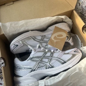 Vita och silverfärgade sneakers - Snygga sneakers i vitt med silverdetaljer och klassisk mesh-design. Skorna har snörning och en sportig look med tydliga linjer på sidan. Perfekta för dig som gillar stilrena och fräscha sneakers till vardags.
