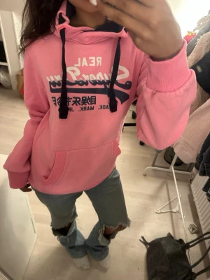Rosa hoodie med tryck och huva - Säljer en rosa hoodie med stor magficka och svart/vita snören i huvan. Tryck i vitt och mörkblått framtill med text och japanska tecken. Hoodien har ribbade muddar och är perfekt för en chill och avslappnad stil.
