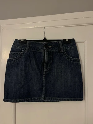 Mörkblå jeanskjol med fickor - Snygg mörkblå jeanskjol i kort modell med klassiska fickor fram och bak. Kjolen har bälteshällor, knapp och dragkedja framtill samt dekorativa fickor med knappar bak. Perfekt till en avslappnad och trendig stil.