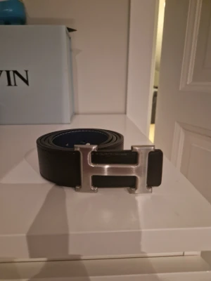 H bälte - Snyggt svart bälte med ett stort, silverfärgat H-format spänne. Bältet är gjort i skinn och har en stilren design som passar till många outfits. Perfekt accessoar för att lyfta din look.