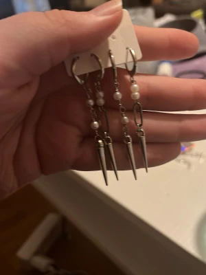 Silvriga örhängen med pärlor och spikes - Säljer ett par statement örhängen i silverfärg med kedjor, små vita pärlor och spetsiga spikes längst ner. Örhängena har en cool och edgy design som sticker ut. Perfekta för dig som vill ha något unikt och trendigt i smyckeslådan.