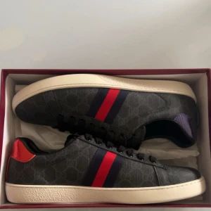 Gucci sneakers med röd och blå detalj - Snygga svarta Gucci sneakers med klassiska röda och blå ränder på sidan. Skorna har en vit sula, svart GG-mönstrad canvas och detaljer i rött och lila metallic på hälen. Perfekta för dig som vill sticka ut med lyxig streetstyle.
