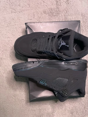 Svarta Air Jordan 4 sneakers - Snygga Air Jordan 4 black cats sneakers med ikonisk Jumpman-logga på plösen. Skorna har meshdetaljer, plastpaneler och en robust sula med synlig luftdämpning. Materialet är en mix av mocka och syntet. Perfekta för dig som gillar streetwear och klassisk sneakerstil.