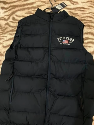 Mörkblå dunväst Polo Club - Mörkblå dunväst från Polo Club med broderad logga på bröstet. Västen har hög krage, dragkedja framtill och två sidofickor med dragkedja. Klassisk quiltad design som håller värmen och passar perfekt till lager-på-lager-stil.