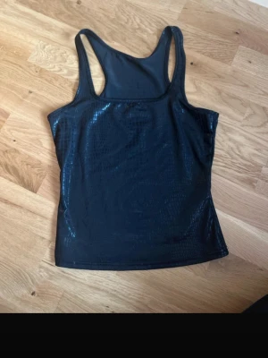Svart glansig linne med ormmönster - Snygg svart linne med glansig finish och ormmönstrad struktur. Modellen är ärmlös med breda axelband och har en tight passform som ger en cool look. Perfekt att styla med jeans eller kjol för en trendig outfit.