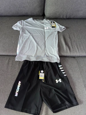 Under Armour träningsset t-shirt & shorts - Snyggt träningsset från Under Armour med en ljusgrå t-shirt och svarta shorts. T-shirten har logga på bröstet och axeln, och shortsen har tryckt logga och text på benet. Båda plaggen är i lätt och snabbtorkande material, perfekt för träning.