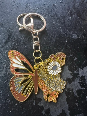 Väsksmycke/Nyckelring fjäril med blommor - Snygg väsksmycke/nyckelring i metall formad som en fjäril med ena vingen dekorerad med blommor i guld, vitt och brunt. Perfekt att hänga på väskan eller nyckelknippan för en unik touch. Färgerna är guld, vit, brun och grå.