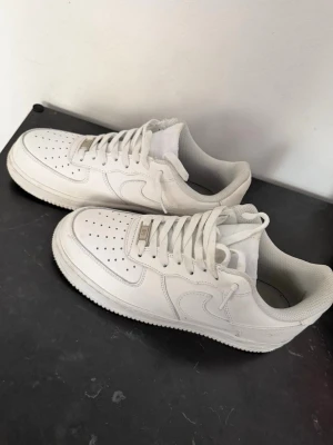Nike Air Force 1 vita sneakers - Klassiska vita Nike Air Force 1 sneakers i lågt utförande. Skorna har en stilren design med perforerad tå, vit sula och Nike-logga på sidan och hälen. Tillverkade i läder med textilfoder och snörning framtill.