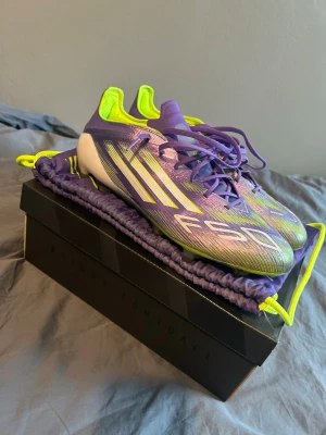 Lila och limegröna Adidas F50 fotbollsskor - Säljer ett par Adidas F50 fotbollsskor i lila och limegrönt med vita detaljer. Skorna har en modern, strömlinjeformad design och är tillverkade i syntetmaterial för lätt vikt och bra grepp på planen. Perfekta för dig som vill sticka ut på fotbollsplanen.