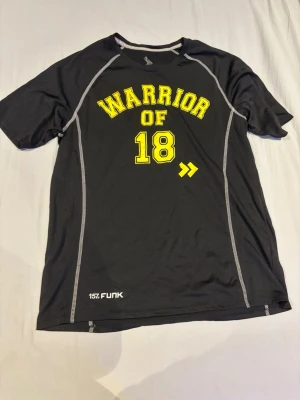 Svart funktions-tshirt WARRIOR OF 18 - Svart funktions-tshirt med trycket 'WARRIOR OF 18' i gul text framtill. T-shirten har vita kontrastsömmar och är tillverkad i ett lätt, syntetiskt material som andas. Perfekt för träning och sportaktiviteter. 
