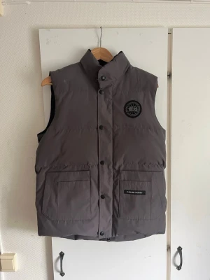 Mörkgrå dunväst från Canada Goose - Snygg mörkgrå dunväst från Canada Goose med hög krage och två stora fickor framtill. Västen har tryckknappar, diskret logga på bröstet och patch på ena fickan. Perfekt för lager-på-lager och håller dig varm under kyliga dagar.