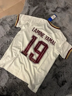 Spanien Lamine Yamal t-shirt Adidas - Vit Spanien t-shirt med Lamine Yamal och nummer 19 på ryggen. Tröjan har vinröda och gula detaljer, korta ärmar och officiella landslagsemblem samt Adidas-logga på bröstet. Tillverkad i lätt och ventilerande polyester, perfekt för fotbollsfans. Storlek M men passar S bra
