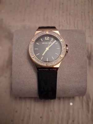 ✨ Michael Kors – Elegant & Lyxig Damklocka ✨ - Stilren klocka från Michael Kors med svart armband och urtavla. Boetten är i guldton och dekorerad med glittrande stenar runt om. Klockan har en enkel och elegant design med tydliga markeringar och MK-logga på armbandet.