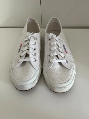 Vita Superga canvas sneakers - Klassiska vita Superga sneakers i canvas med lågt skaft och vita snören. Skorna har en vit gummisula och Superga-logga på sidan och hälen. 