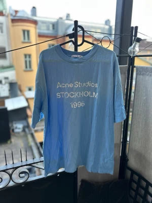 Blå Acne Studios t-shirt - Säljer en ljusblå t-shirt från Acne Studios med trycket 'Acne Studios STOCKHOLM 1996' på bröstet. Klassisk rund hals och korta ärmar. Perfekt för dig som gillar stilrena och trendiga plagg.