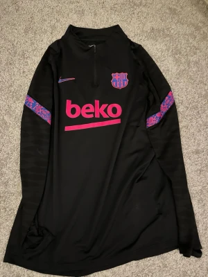 Barcelona tröja - Svart långärmad träningströja från FC Barcelona med rosa och lila detaljer samt klubbmärke på bröstet. Tröjan har half zip och mönstrade band på ärmarna. Perfekt för fotbollsträning eller sportiga aktiviteter.