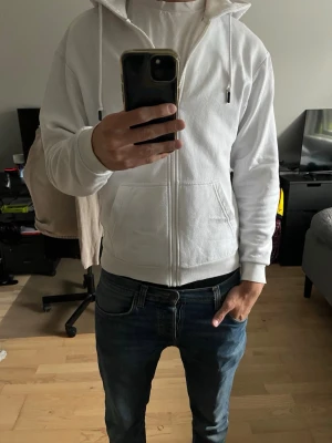 Vit zip hoodie med huva - Säljer en clean vit hoodie med dragkedja och stor huva. Hoodien har två fickor framtill och ribbade muddar vid ärmar och midja. Perfekt för en avslappnad och stilren look. Passar till det mesta och är riktigt skön att bära. Nopprig!