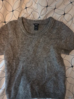 Grå stickad tröja från Lindex - Mohair - Mysig grå stickad tröja från Lindex med korta ärmar och rund halsringning. Tröjan har en enkel och klassisk design med ribbstickade muddar vid ärmslut och nederkant. Perfekt att matcha med jeans eller kjol för en avslappnad look.