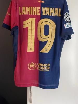 Barcelona matchtröja Lamine Yamal - Säljer en officiell FC Barcelona matchtröja från Nike med Lamine Yamal och nummer 19 på ryggen. Tröjan är röd och blå med guldfärgade detaljer, klubbmärke och sponsorer på bröstet. Tillverkad i lätt och ventilerande material, perfekt för fotbollsträning eller match.