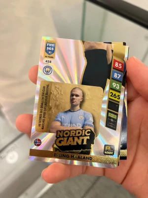 Samlarbild Nordic Giant Erling Haaland - Säljer en glittrig samlarbild från FIFA 365-serien med Erling Haaland i Manchester City-tröja. Kortet har holografisk yta, guldiga detaljer och visar statistik samt texten 'Nordic Giant'. Perfekt för fotbollsfans och samlare.