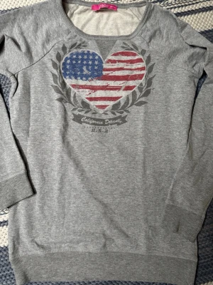 Grå sweatshirt med USA-hjärta - Grå sweatshirt med tryck av ett hjärta i amerikanska flaggans färger och texten 'California Dream U.S.A.' framtill. Tröjan har rund halsringning, långa ärmar och ribbade muddar vid ärmslut och nederkant. Tillverkad i mjukt bomullsmaterial.storlek 158/164 o passat st xs/s